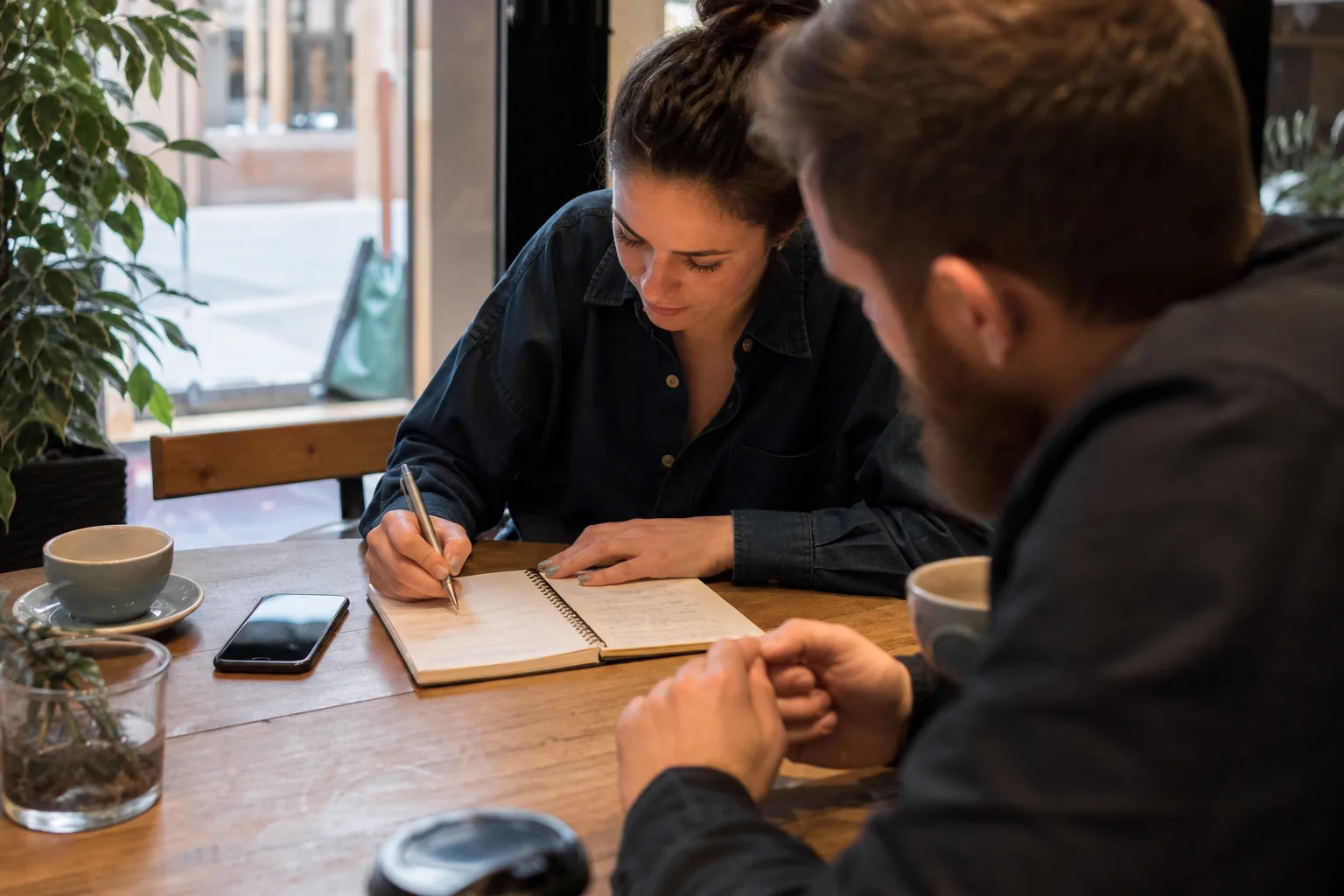 Two adults at a cafe table comparing name ideas in a paper notebook next to a phone